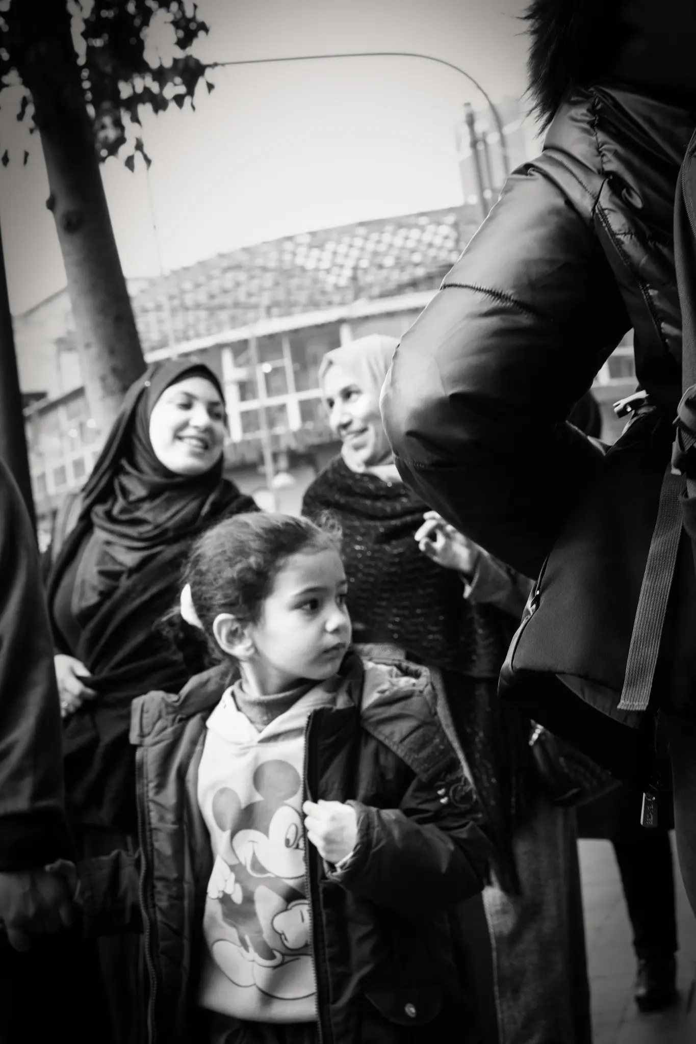 Una bambina osserva i passanti mentre due donne sorridono alle sue spalle.