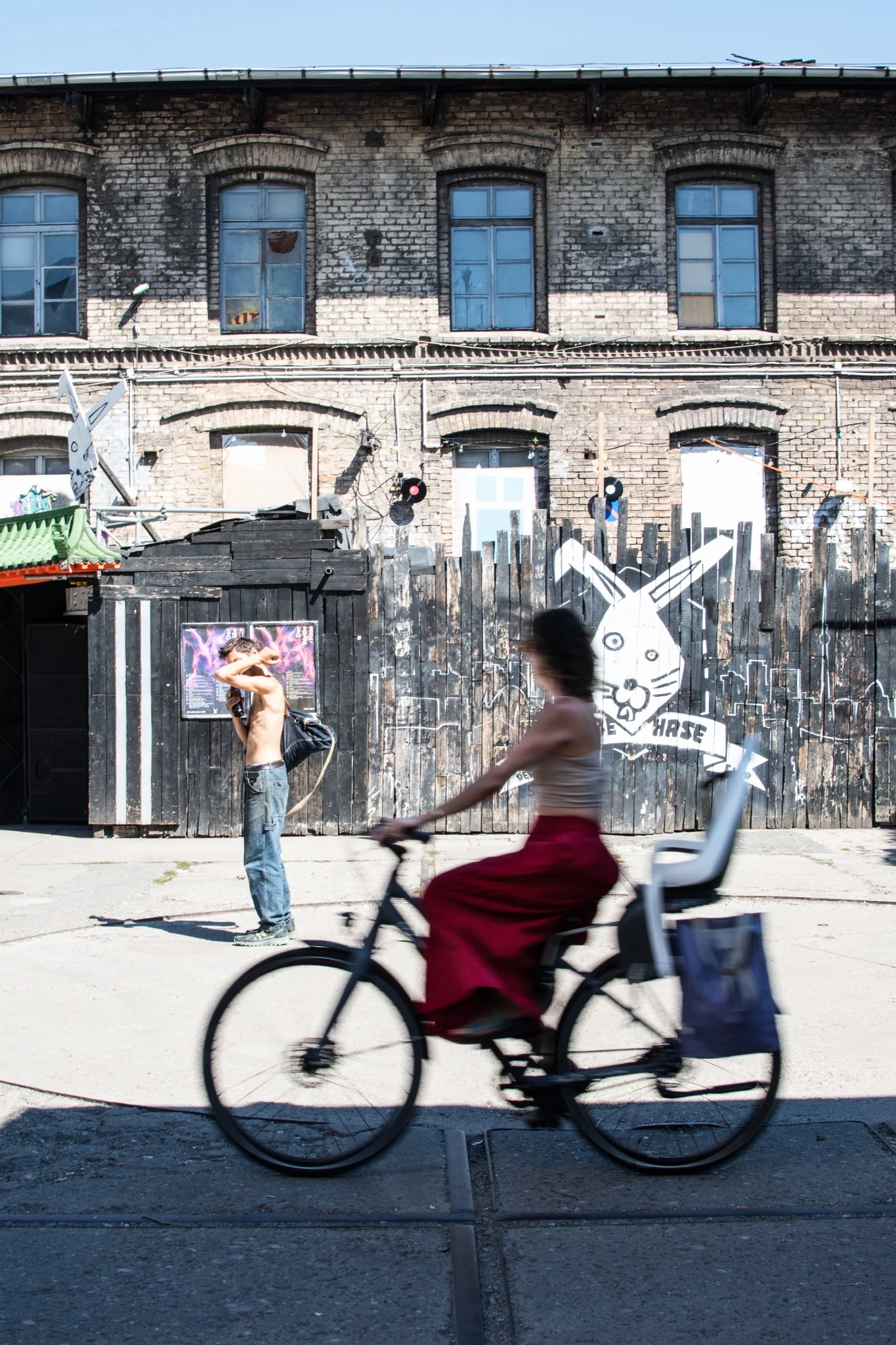 A woman on a bicycle crosses the scene in front of a painted fence while a man shields his face from the sun in Berlin.